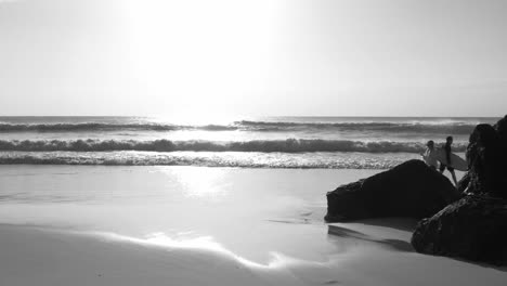 black and white -  surfer with surfboard walking on the beach shore with bright sky and ocean waves in the background - palm beach in gold coast, qld, australia - wide slowmo shot