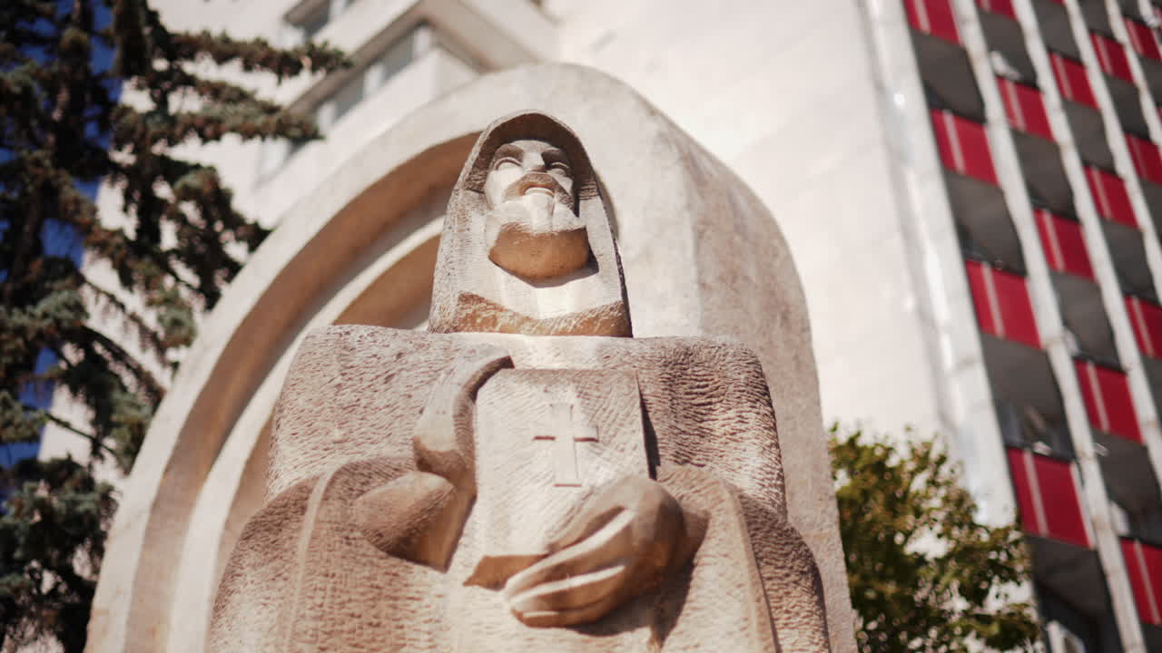 Low angle view of a carved stone statue Petru Movila holding a book with a cross, framed by trees and modern architecture, Chisinau, Moldova