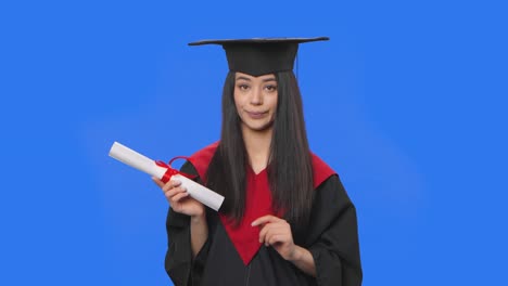 retrato de una estudiante en traje de graduación de gorra y túnica, sosteniendo un diploma y mostrando el pulgar hacia abajo. mujer joven posando en el estudio con fondo de pantalla azul. de cerca. cámara lenta lista 59.94fps