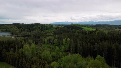 Scenic-4K-aerial-drone-flight-with-view-upon-Bavaria's-idyllic-lakes-in-the-rural-countryside-with-a-beautiful-sky-and-the-alps-mountains-in-the-background,-green-lush-fields-and-forest-in-spring-time
