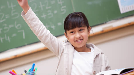 smiling asian elementary schoolgirl raising hand sitting in school classroom