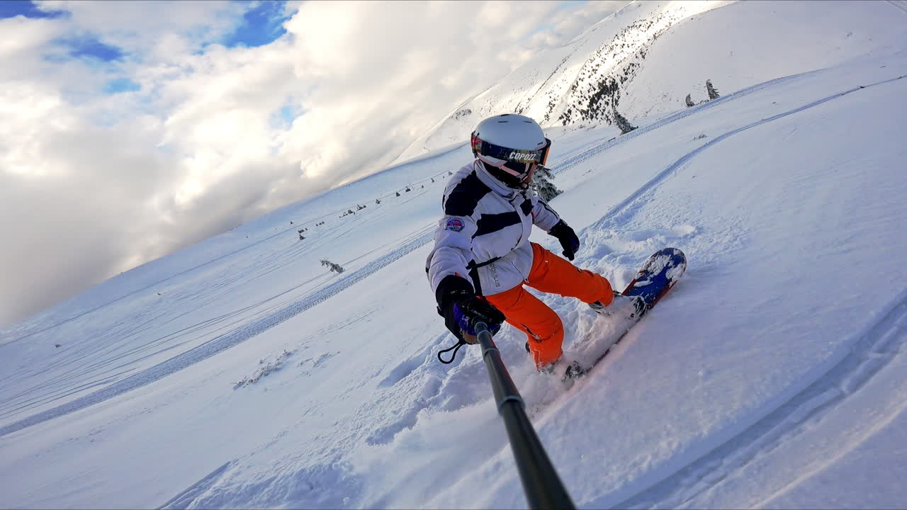 Snowboarder rides by the snowy mountain slope taking selfie video. Amazing scenery at backdrop.