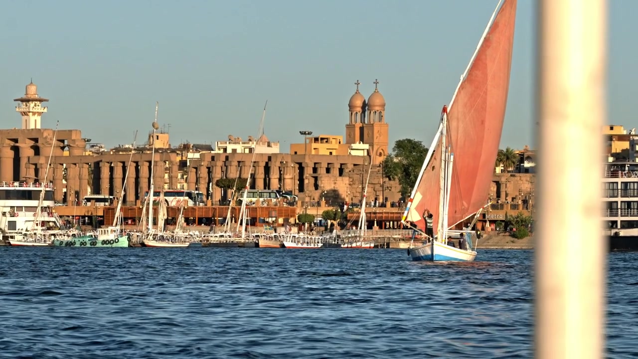 Un'ampia vista al tramonto che guarda attraverso le acque blu del fiume Nilo verso la riva di Luxor, fiancheggiata da feluche ormeggiate, le antiche colonne del Tempio di Luxor e importanti chiese cristiane.