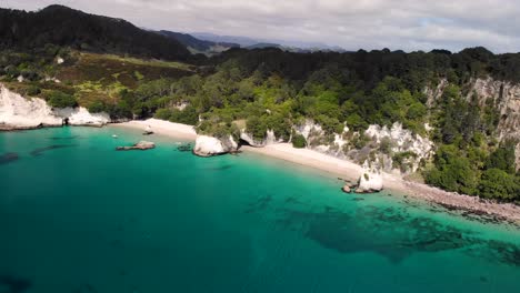 beautiful white sandy beach throught tunnel cave surrounded limestone cliffs and forested hills at new zealand seaside aerial orbit reveal shot