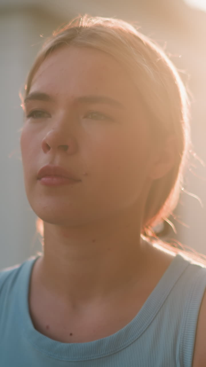 젊은 여자의 가까운 모습은 사려 깊게 보이고, 빛이 그녀의 얼굴을 따뜻한 빛으로 비추고, 롤러 스케이팅처럼 우아하게 움직이고, 흐릿한 배경에서 건물과 나무의 부분적인 시야입니다.