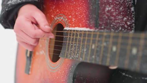 close-up of hand strumming acoustic guitar strings dusted with snow, showcasing vibrant red-orange guitar tones and frosted details, a perfect harmony between music and winter's