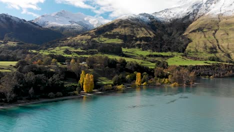 slowmo - flying over beautiful blue lake wakatipu, queenstown, new zealand with mountains fresh snow, clouds, blue sky autumn trees in the background - aerial drone