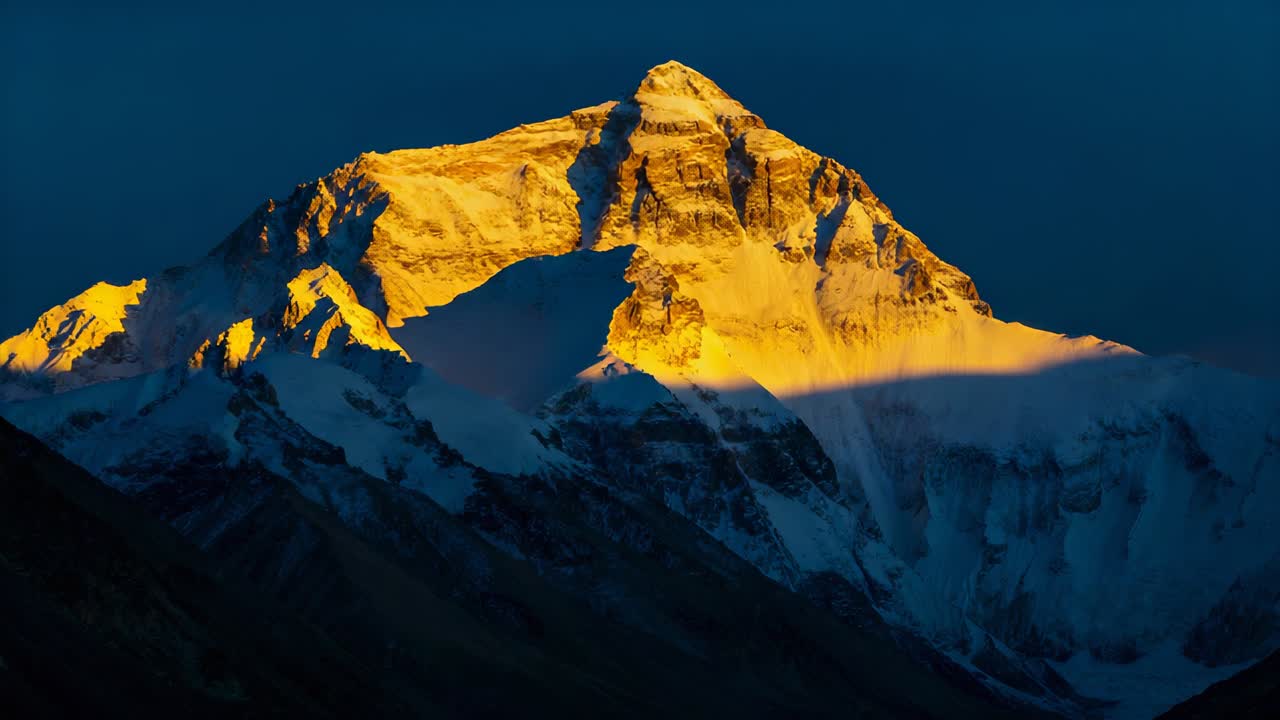 Vetta montuosa innevata immersa nella luce ad alta quota, alpenglow dell'alba che si diffonde e fascia di nuvole che si dirada.