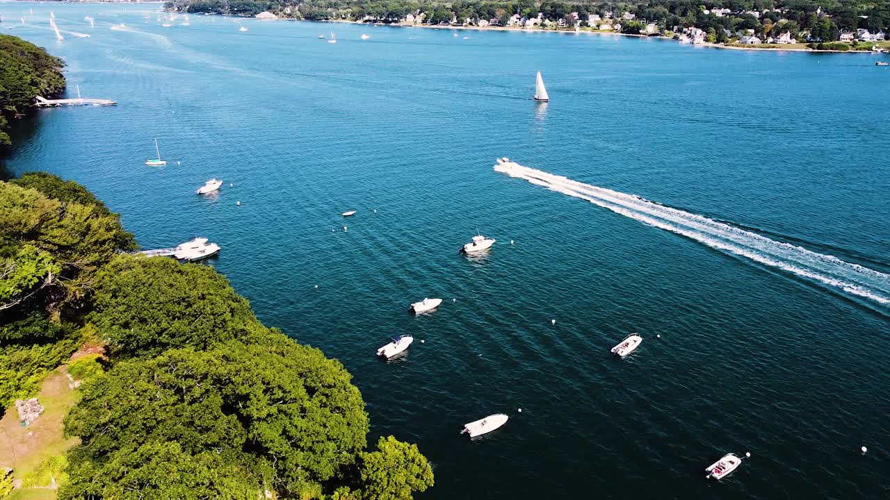 Aerial shot of island home flying towards water with sailboats and speedboat crossing open water near Portland Maine
