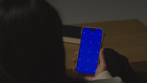 Over-The-Shoulder-Shot-Of-Woman-At-Home-Sitting-On-Sofa-With-Blue-Screen-Mobile-Phone-Scrolling-Through-Internet-Or-Social-Media-2