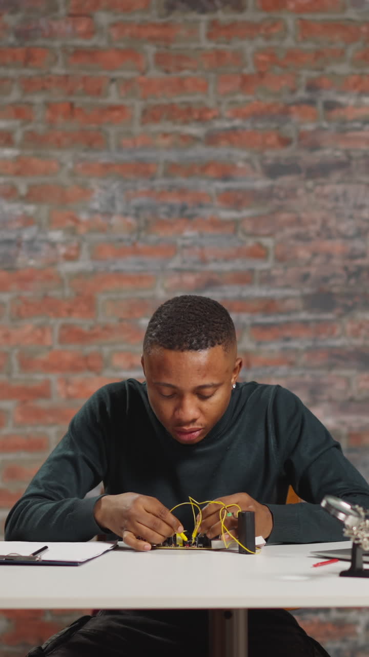 Young African-American engineer man tries unfortunately to assemble electrical device panel sitting at workplace in scientific laboratory premise