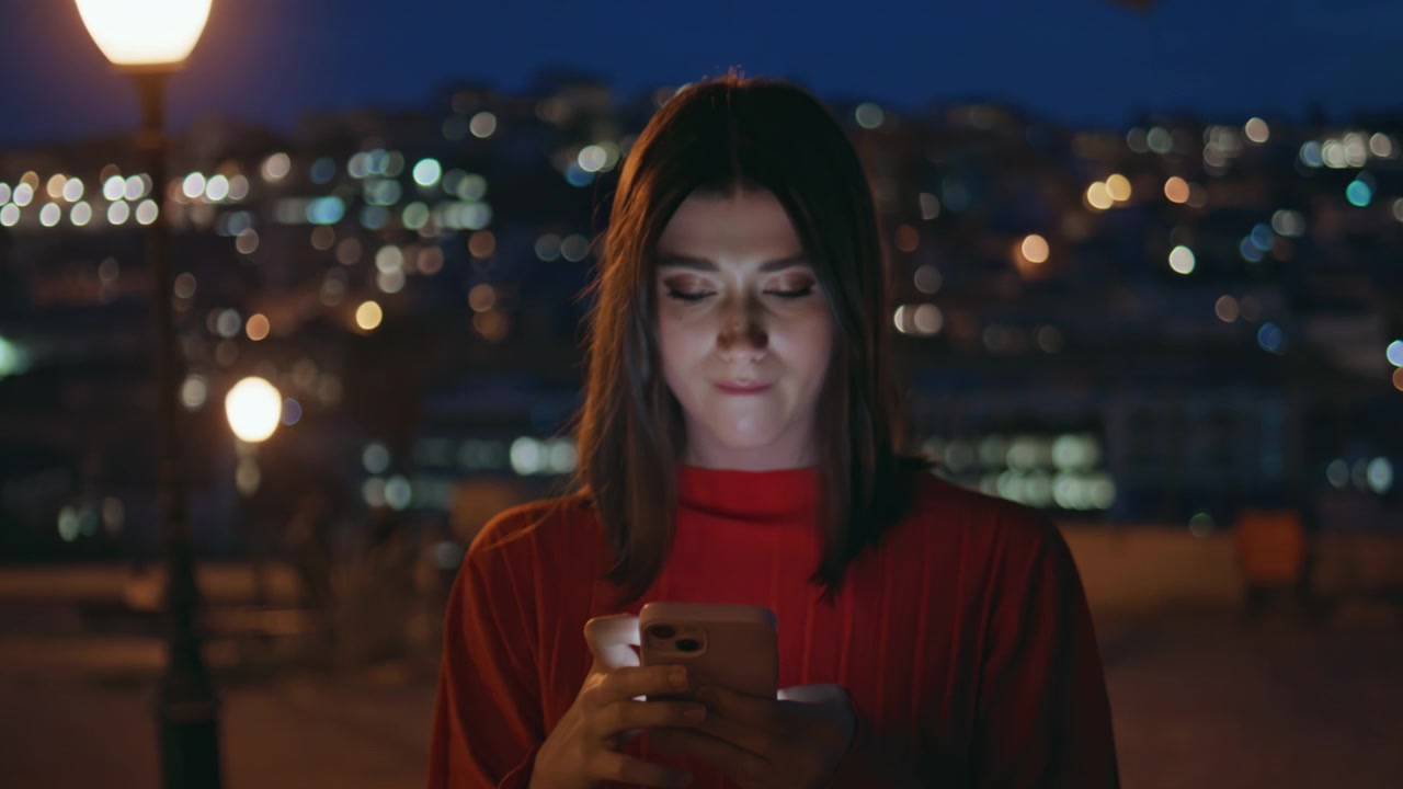 Giovane donna che scorre lo smartphone in una strada cittadina di notte. Primo piano mentre messaggia di sera.