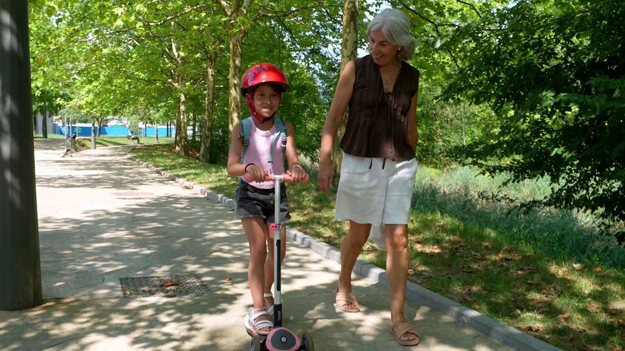 Grandmother Walking With Granddaughter Riding Scooter in Park