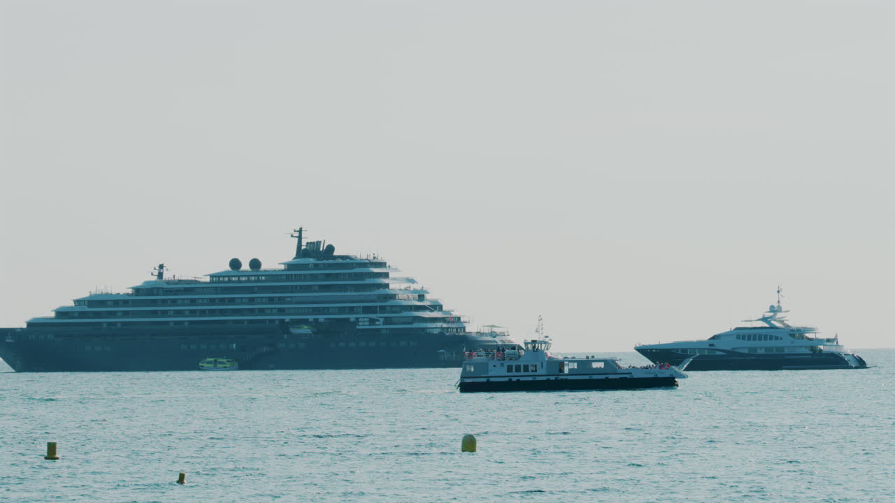 Large cruise ship and luxury yachts anchored on calm blue sea under hazy sky