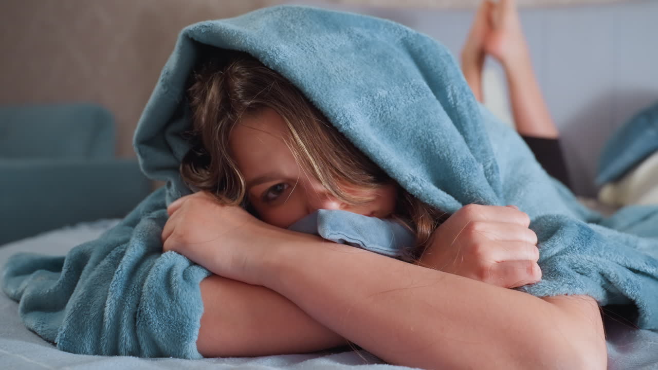 Primo piano di una giovane ragazza avvolta in una morbida coperta blu con il cappuccio sulla testa, che appoggia le braccia sul letto mentre guarda serenamente avanti, evocando calore, quiete e comfort in un tranquillo ambiente da camera da letto al chiuso.