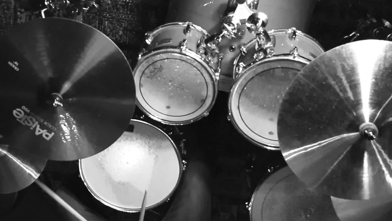 Grayscale top down footage of a person with tattoos playing and hitting acoustic drums in slow motion in a studio room. Man hits and plays cymbals, crashes, snare drum, tom toms and even bass drum.
