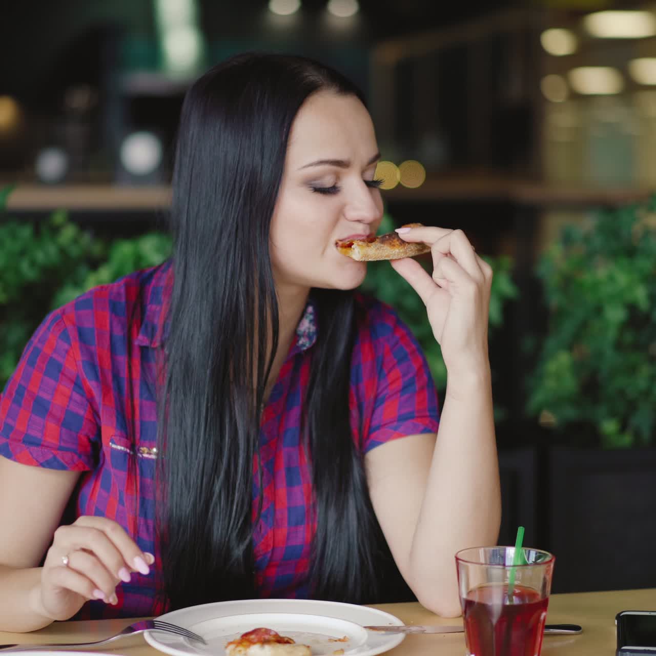 beautiful girl in a plaid shirt is eating a slice of pizza at a table in the pizzeria