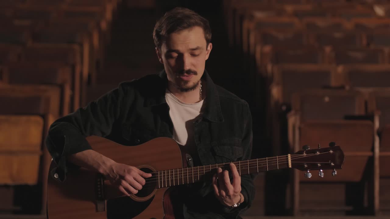un chico guapo toca una guitarra acústica en un cine abandonado. el músico canta una canción y acompaña en la guitarra