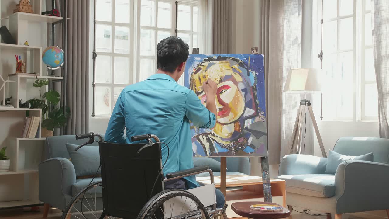 Hind View Of An Asian Artist Man In Wheelchair Holding Paintbrush Mixed Colour And Painting A Girl'S Face On The Canvas