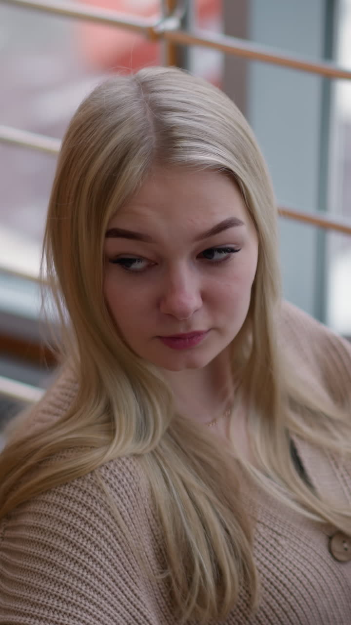 긴 금발 머리카락을 가진 우아한 여성의 클로즈업 현대 쇼핑몰에 앉아, 사려 깊게 쳐다보고, 큰 창문으로 보이는 도시 풍경과 흐릿한 눈 인 거리를