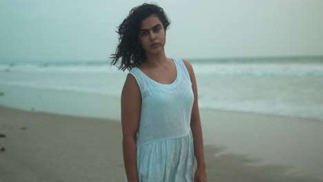 woman in white dress stands contemplatively on a cloudy beach at dusk, waves gently breaking