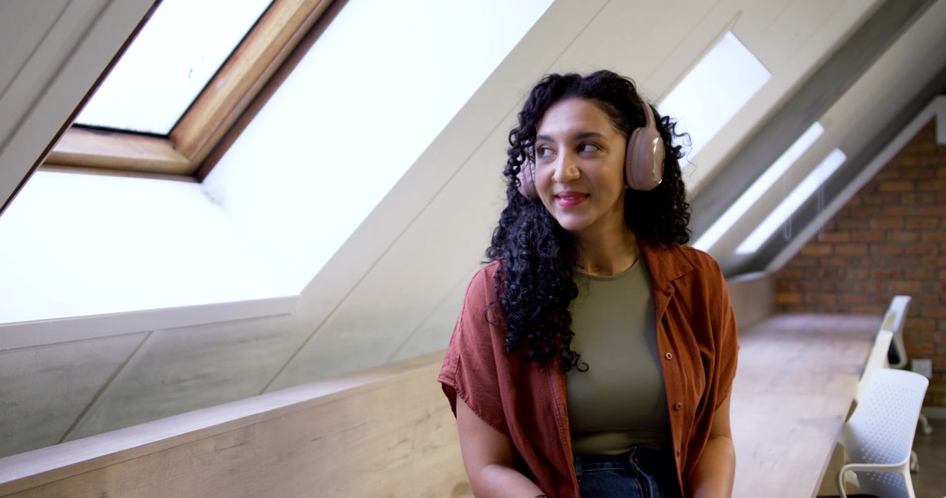 Woman sitting on bench in shirt, pink headphones, noticing skylight and turning, observing outside