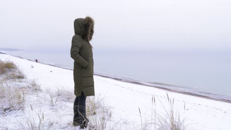 Mujer-Joven-Con-Ropa-De-Invierno-Verde-Se-Encuentra-En-La-Orilla-Arenosa-De-La-Playa-Del-Mar-Báltico,-Hierba-Nevada,-Foque-Medio-Disparado