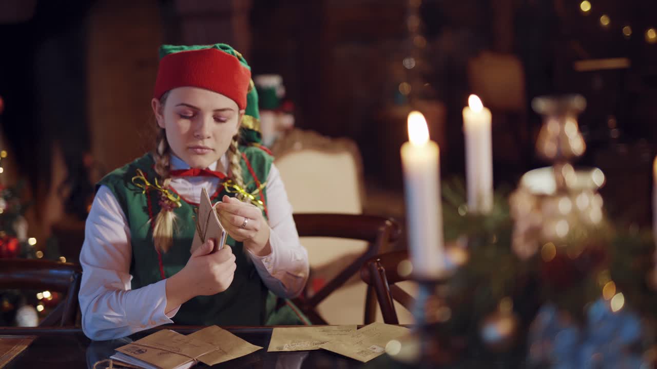 Happy female elf holding a stack of envelopes. Funny elf sitting at the table and looking through the envelopes for Santa Claus in the Christmas decorated room.