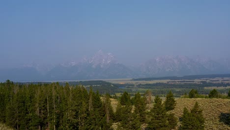 Drone-Aéreo-Paisaje-Tiro-Natural-Pasando-Sobre-Las-Copas-De-Los-Pinos-Con-La-Cordillera-Del-Parque-Nacional-Grand-Tetons-Con-Un-Valle-De-Arbustos-Y-Pinos-Detrás-En-Wyoming,-Estados-Unidos