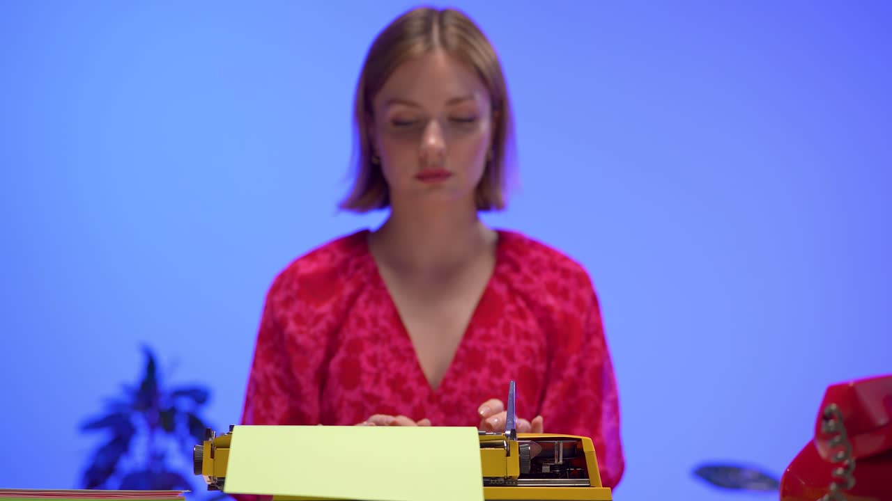 A calm woman in a vibrant red floral dress types on a yellow retro typewriter. Set in a minimal blue workspace, this scene blends vintage aesthetics with focused, analog creativity