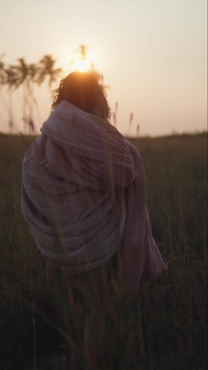 Vertical footage of a woman strolling outdoors at sunrise or sunset, blanket draped around her