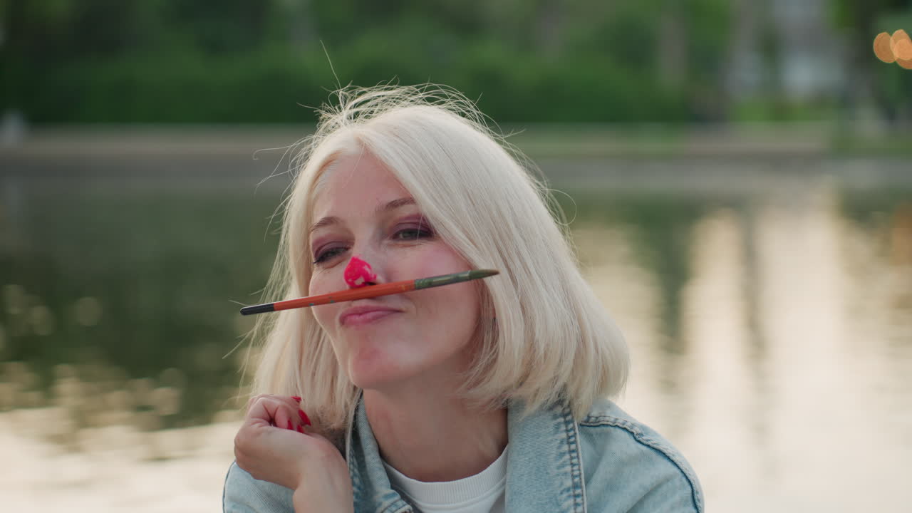close up joyful woman with paint on nose balancing brush on nose while seated by calm water at sunset in blurred park, playful art moment capturing creativity and casual fun outdoors