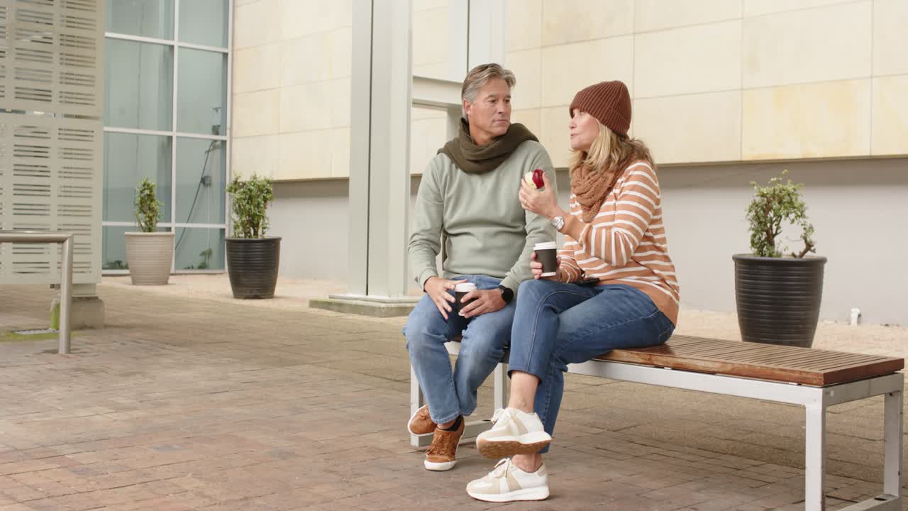 Una pareja mayor señala hacia arriba mientras come una manzana y toma café para llevar en un banco en el patio.