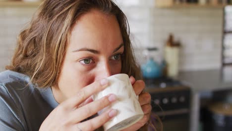 Thoughtful-caucasian-woman-in-sunny-cottage-kitchen-drinking-coffee