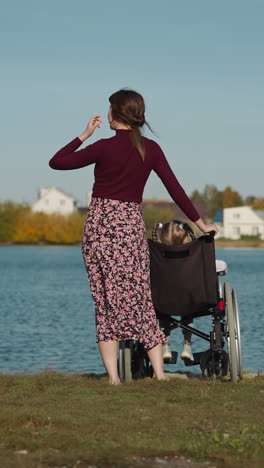 kid with disability spends time with mother. concentrated woman straightens hair standing near little girl in wheelchair. family rests on river bank on sunny day backside view