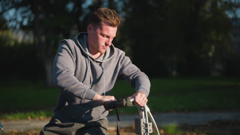 guy in sitting position wearing gray long-sleeved shirt, focusing on fixing a stilt. background is blurred, highlighting his concentrated expression and detailed stilt adjustment in outdoor setting