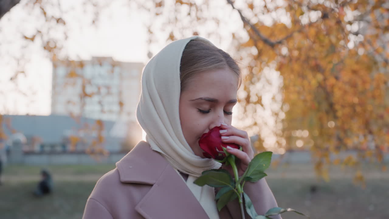 Молодая белая женщина в платке, поэтесса рассматривает лепесток розы, студентка, вдыхающая аромат цветка, модель, улыбающаяся в камеру, вечерний парк с золотыми листьями берёзы, мягкий закатный контровой свет, городские здания вдалеке, нежно.