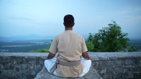 unrecognizable indian male doing hatha yoga and meditating on top of a hill on castle wall at sunrise shot from behind overlooking the valley bellow in traditional yogi clothes