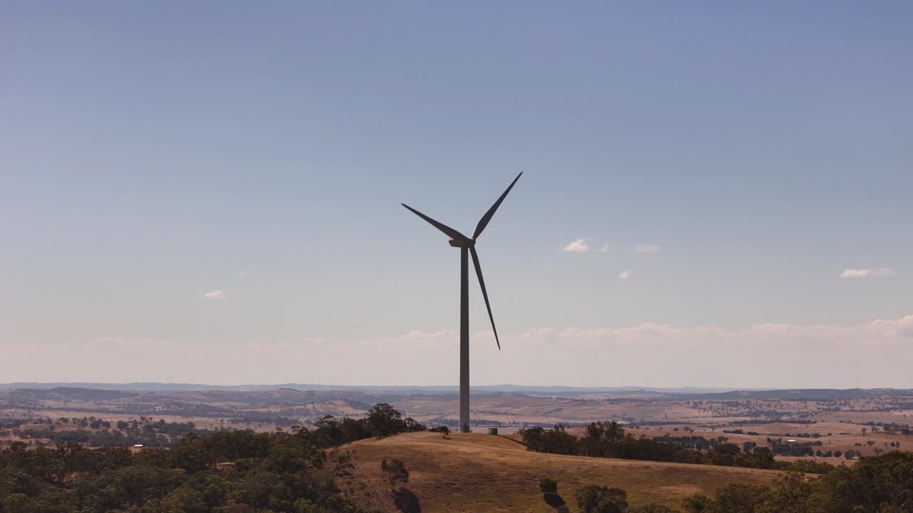 Ampia ripresa aerea orbitale di una singola turbina eolica isolata, su uno sfondo di campagna pianeggiante e cielo azzurro chiaro