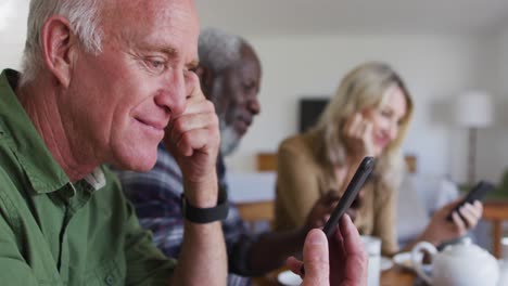 two diverse senior couples sitting by a table drinking tea using smartphones at home