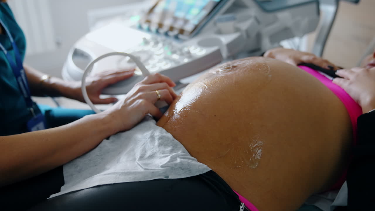 Pregnant woman is examined on ultrasonic equipment. Female doctor's hand moves the device by the big gelled belly of a future mom. Baby is kicking. Close up.