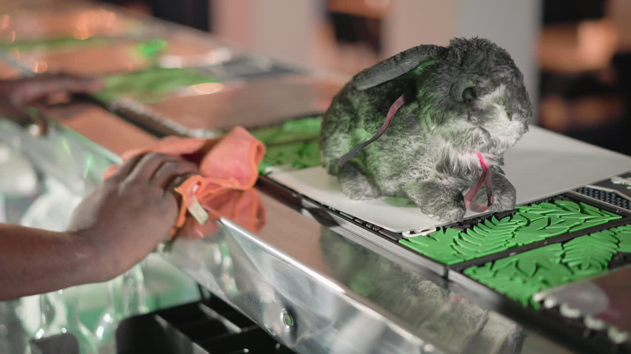 Close-up of bartender cleaning bar surface with orange cloth, with plush rabbit on counter. Green Heineken neon sign glowing in background. Detailed focus on cleaning task and bar decor