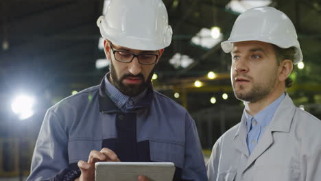 two workers wearing helmets talking while looking at the tablet in a big factory