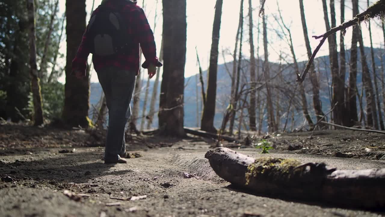 Aufnahme aus der Froschperspektive eines Mannes, der am Sandstrand entlanggeht, im Regenwald der Westküste Kanadas mit Bergen im Hintergrund