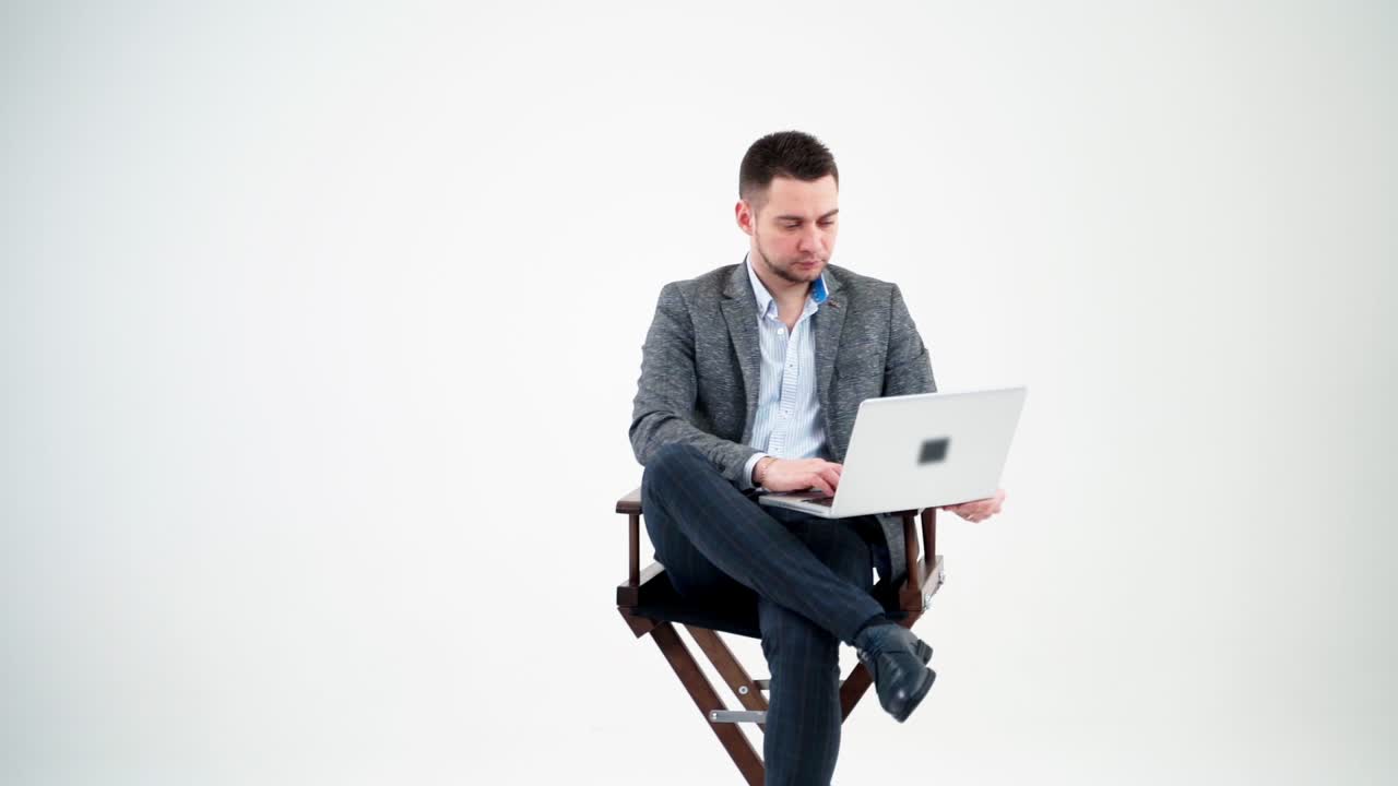 Young man sitting in a chair with laptop. Handsome businessman in stylish costume working on a modern computer, isolated in studio. Freelance work.