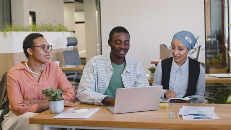 young worker working with laptop sitting at his desk while muslim businesswoman and businesswoman talk to him sitting at desk 1