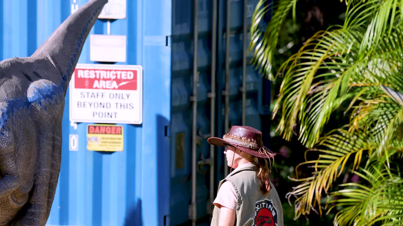 A performer in ranger attire interacts with a realistic velociraptor puppet outdoors, beside blue shipping containers and lush greenery, under bright natural sunlight