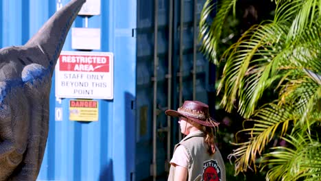 a performer in ranger attire interacts with a realistic velociraptor puppet outdoors, beside blue shipping containers and lush greenery, under bright natural sunlight