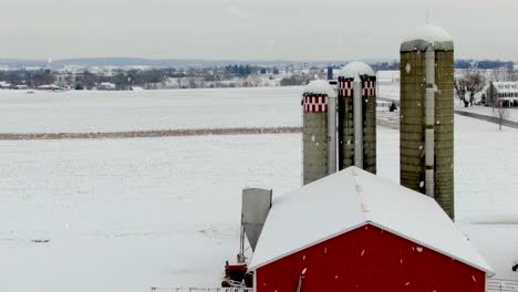 reverse aerial reveals red barns and farm during winter snowstorm