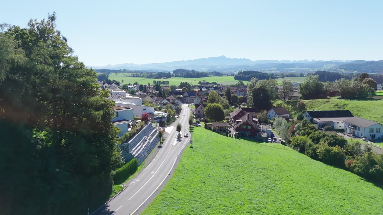 Town emerges beyond forested slopes as road curves into valley, Swiss countryside; drone reveal